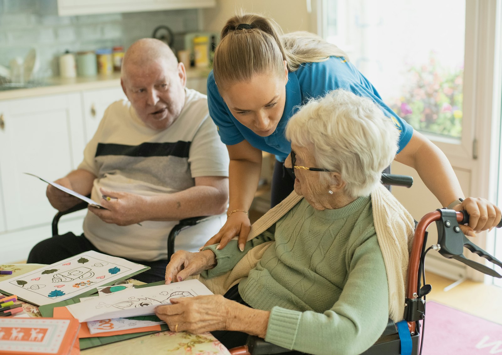 A caregiver leans in to help an older woman in a wheelchair with a coloring activity at a table (stock photo)